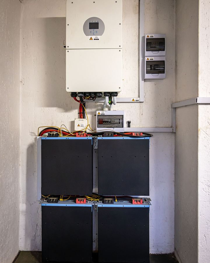 A wall-mounted hybrid inverter above four stacked battery modules in a plant room